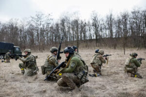 A group of Ukrainian soldiers in camouflage uniforms and helmets, kneeling and holding rifles, conduct a training exercise in an open field with sparse grass and leafless trees in the background. A military truck is partially visible on the left side.
