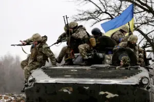 A group of Ukrainian soldiers in camouflage uniforms stand atop a tank in a snowy forest, holding rifles and displaying the blue and yellow Ukrainian flag emblem, with winter trees and terrain in the background.