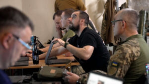 A group of military personnel in uniform gathered around a conference table in a command center, engaged in a strategic briefing with laptops and documents.