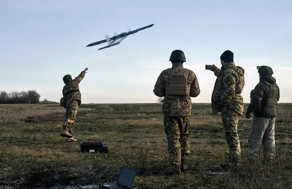 A group of Ukrainian soldiers in camouflage uniforms and helmets operate a drone in an open grassy field, with one using a laptop for control as the UAV ascends against a soft horizon line at dawn.
