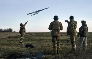 A group of Ukrainian soldiers in camouflage uniforms and helmets operate a drone in an open grassy field, with one using a laptop for control as the UAV ascends against a soft horizon line at dawn.