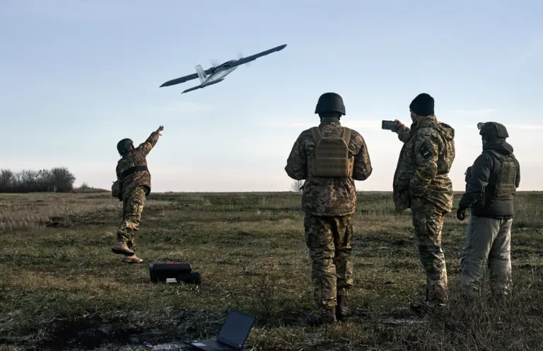 A group of Ukrainian soldiers in camouflage uniforms and helmets operate a drone in an open grassy field, with one using a laptop for control as the UAV ascends against a soft horizon line at dawn.