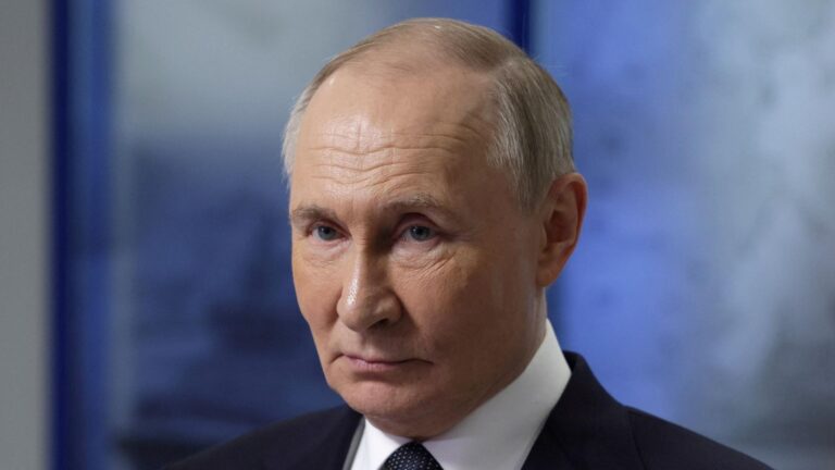 A close-up image of Vladimir Putin, the Russian President, with a serious expression, wearing a dark suit and tie, against a blurred blue background.