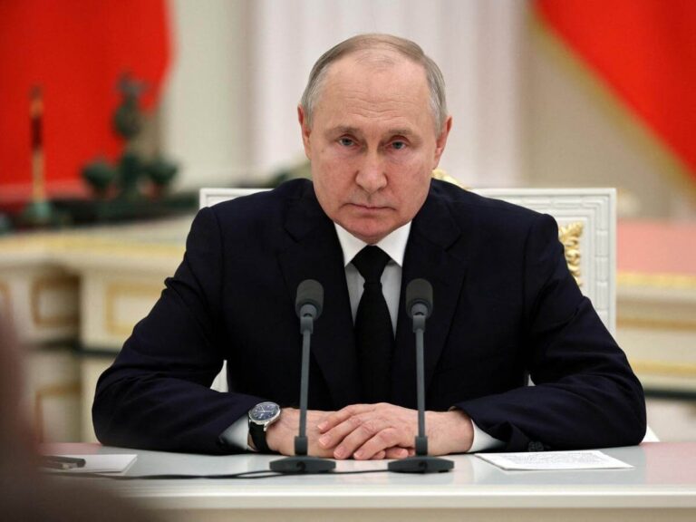 A person in a dark suit and tie sitting at a table with microphones, hands clasped, against a backdrop of red and white decor.