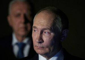 A close-up image of Vladimir Putin with a serious expression, illuminated by soft lighting against a dark background, with a blurred figure in a suit and tie standing behind him.