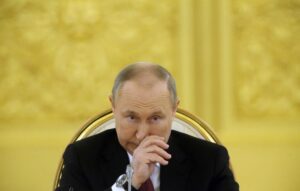 Vladimir Putin, in a black suit and red tie, sits in an ornate golden chair with a thoughtful expression, hand near his face, against a richly decorated yellow background.