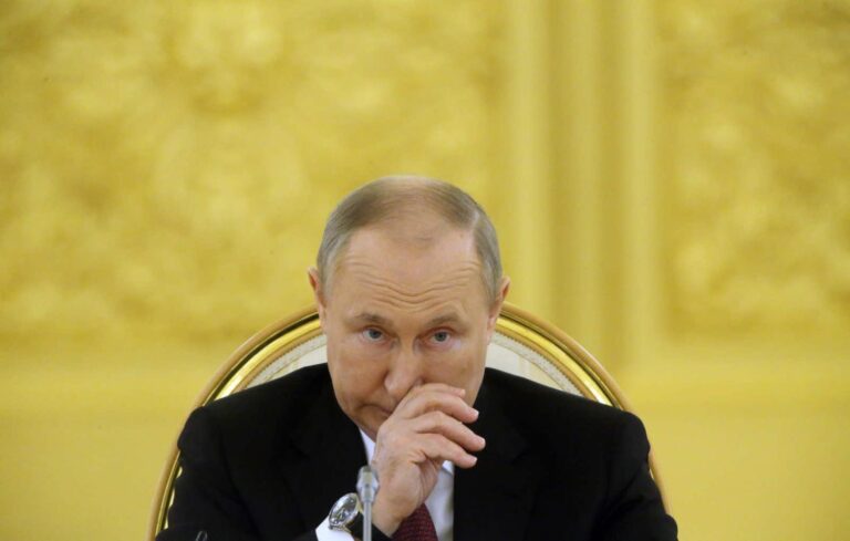 Vladimir Putin, in a black suit and red tie, sits in an ornate golden chair with a thoughtful expression, hand near his face, against a richly decorated yellow background.