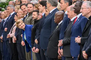 A group of world leaders in formal attire standing in a line, with Volodymyr Zelenskyy prominently leaning forward, smiling, among other notable figures in suits and ties, symbolizing international solidarity.