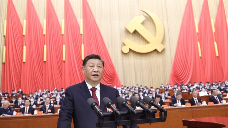 Xi Jinping speaking at a podium with multiple microphones, standing in front of a large audience and a backdrop featuring the hammer and sickle symbol, surrounded by red curtains.