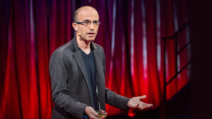 Yuval Noah Harari speaking on stage about AI and human evolution, with a thoughtful expression against a backdrop of futuristic elements.