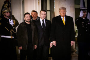 A formal gathering outside a grand building, featuring three prominent leaders—Volodymyr Zelensky, Emmanuel Macron, and Donald Trump—flanked by ceremonial guards in ornate uniforms saluting, with flags and columns in the background.