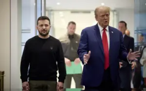 Image of U.S. President Donald Trump in a blue suit with a red tie gesturing with both hands, walking alongside Ukrainian President Volodymyr Zelensky, who wears a black sweater with a Ukrainian emblem. They are in a modern corridor with blurred security personnel and officials in the background, indicating a formal diplomatic meeting.