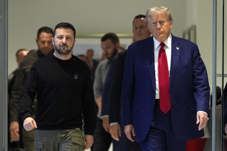 A photo featuring two prominent figures walking side by side in a formal setting, with one dressed in a dark sweater and cargo pants and the other in a blue suit with a red tie, accompanied by a group of people in the background.