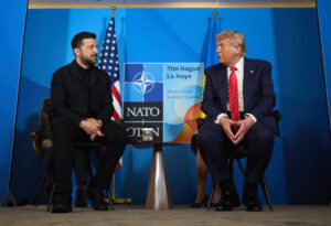 Two leaders, Volodymyr Zelensky in a dark suit and Donald Trump in a blue suit with a red tie, seated side by side at a NATO Summit in The Hague, with the NATO logo and flags of the U.S. and Ukraine in the background.