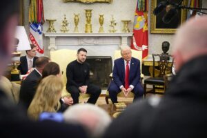 Photograph of President Donald Trump and Ukrainian President Volodymyr Zelenskyy seated in the Oval Office, engaged in a meeting with microphones and aides present. The background features a fireplace with golden trophies, military flags including the U.S. Army and Marine Corps, and a bust, with the presidential seal on a box in the foreground. Taken on July 13, 2025, highlighting a key diplomatic moment.