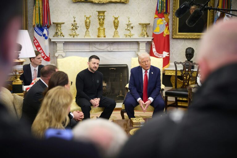 Photograph of President Donald Trump and Ukrainian President Volodymyr Zelenskyy seated in the Oval Office, engaged in a meeting with microphones and aides present. The background features a fireplace with golden trophies, military flags including the U.S. Army and Marine Corps, and a bust, with the presidential seal on a box in the foreground. Taken on July 13, 2025, highlighting a key diplomatic moment.