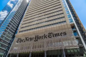 A tall modern building with the words "The New York Times" prominently displayed on its facade, surrounded by other high-rise structures under a bright blue sky.