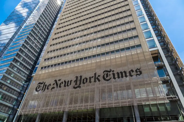 A tall modern building with the words "The New York Times" prominently displayed on its facade, surrounded by other high-rise structures under a bright blue sky.